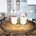 Load image into Gallery viewer, The Boho Bliss: Custom Round Serving Tray for Effortless Entertaining with ornate carvings and black handles holds two white teacups on a rustic wooden surface against a blurred kitchen backdrop.
