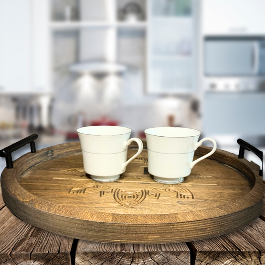 The Boho Bliss: Custom Round Serving Tray for Effortless Entertaining with ornate carvings and black handles holds two white teacups on a rustic wooden surface against a blurred kitchen backdrop.