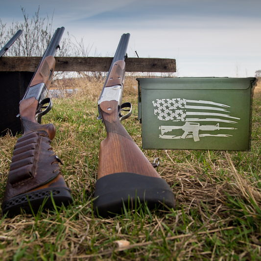 Two shotguns rest on the grass near a Tactical Titan: Personalized Military Style Storage Can, featuring a white U.S. flag stencil and AR-15. A wooden bench and dry vegetation are in the background under a cloudy sky, evoking a rugged outdoor setting.