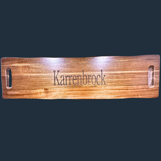 The Sophisticated Dining: Engraved Grazing Board is a rectangular acacia wood tray featuring a smooth, polished surface and cutout handles on each side, with Karrenbrock engraved in the center, making it an elegant and effortless entertaining accessory.
